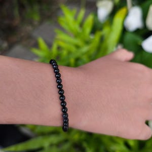 Puede incluir: Una pulsera de cuentas negras se lleva en la muñeca. La pulsera está hecha de pequeñas cuentas negras redondas y pulidas. El fondo está borroso, con follaje verde visible. La pulsera probablemente está hecha de piedras preciosas.