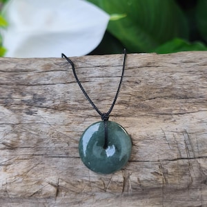 Puede incluir: Un collar con colgante de jade verde oscuro con una piedra circular en forma de rosquilla. La piedra cuelga de un cordón negro, sobre un fondo de madera envejecida. El colgante mide aproximadamente 3,8 cm de diámetro.