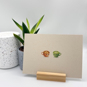 Könnte beinhalten: Eine Grußkarte mit zwei Aquarell-Kaffeetassen mit lächelnden Gesichtern. Eine Tasse ist orange mit einem Herzmuster, die andere ist grün mit einem Blättermuster. Die Karte wird von einem Holzständer gehalten.