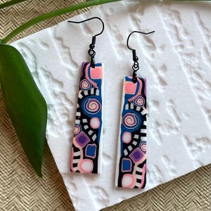May include: A pair of dangle earrings featuring long, rectangular pendants with a vibrant abstract pattern of pink, purple, blue, black, and white shapes, including swirls and squares, suspended from black metal ear wires.