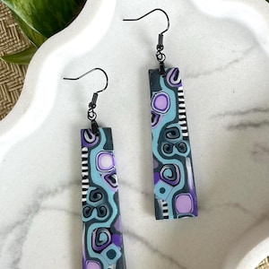 May include: A pair of long, rectangular dangle earrings featuring a complex pattern of purple, light blue, dark gray, and white shapes. The earrings are suspended from black metal fishhook wires and display a mix of circular and linear motifs.