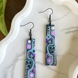 May include: A pair of rectangular dangle earrings with black hooks. The earrings feature a gray base with a pattern of light blue, purple, and white shapes and swirls. The earrings are displayed on a white, wavy-edged tray.