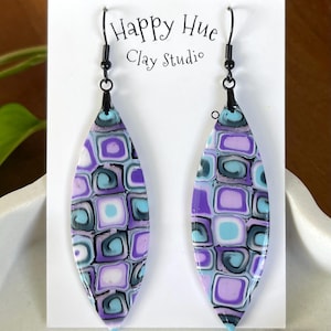 May include: A pair of handmade, long, teardrop-shaped earrings. The earrings feature a mosaic pattern in shades of purple, blue, and white. The text "Happy Hue Clay Studio" is visible on the white card behind the earrings. The earrings have black metal hooks.