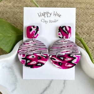 May include: Dangle earrings featuring a square top piece and a larger circular bottom piece, both with a marbled pattern in hot pink, black, and white. The earrings are displayed on a white card labeled Happy Hue Clay Studio.