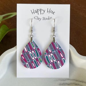 May include: Teardrop-shaped earrings with a colorful pattern of magenta, teal, white, and gray. The earrings are displayed on a white card with the text "Happy Hue Clay Studio". The earrings have silver-colored hooks.
