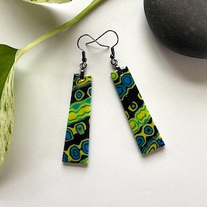 May include: A pair of dangle earrings featuring long, rectangular pendants with a vibrant, multi-colored abstract pattern in shades of lime green, bright blue, and black. The earrings are attached to thin, black metal ear wires for hanging.