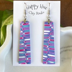 May include: A pair of rectangular dangle earrings with a colorful pattern. The earrings feature a design of horizontal lines in shades of purple, pink, blue, and white. The earrings are hanging from silver-colored hooks. The card behind the earrings says "Happy Hue Clay Studio."