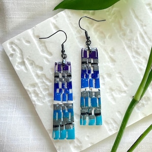 May include: A pair of rectangular drop earrings featuring a mosaic pattern of blue, purple, silver, and white segments. The earrings hang from black metal fishhook wires and have a flat, smooth finish with a gradient color transition.