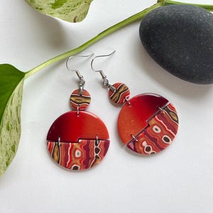 May include: Dangle earrings featuring a small circular stud connected to a larger circular drop. Each piece combines a solid, shimmering red upper half with a lower half displaying a multi-colored, wavy, agate-like pattern in shades of red, brown, and white.