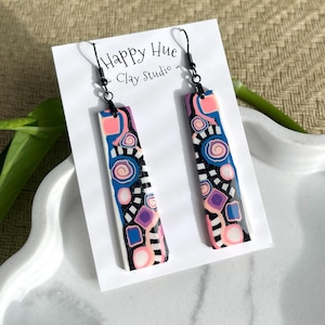 May include: A pair of long, rectangular dangle earrings featuring a black base with abstract patterns in blue, pink, purple, and white. The earrings hang from black metal hooks on a white display card labeled Happy Hue Clay Studio.