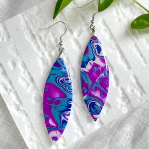 May include: A pair of marquise-shaped dangle earrings featuring a vibrant marbled pattern in shades of bright magenta, electric blue, and white. The earrings are suspended from silver-toned metal hooks and have a smooth, glossy finish on the surface.