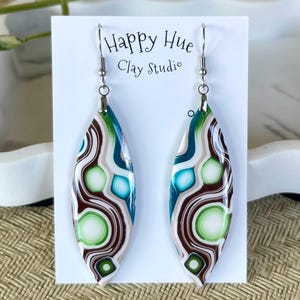 May include: A pair of leaf-shaped earrings with a marbled design. The earrings feature a pattern of green, brown, blue, and white swirls. The text "Happy Hue Clay Studio" is printed on the white card behind the earrings.