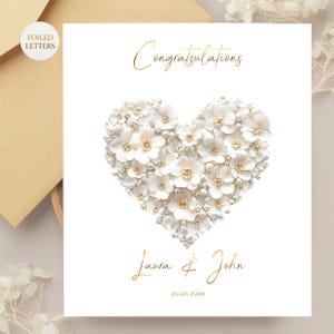 A white greeting card with a heart shape made of small white flowers and pearls, with gold foil text reading "Congratulations" above and "Laura & John 20.05.2026" below, on a light brown envelope and a blurred background.