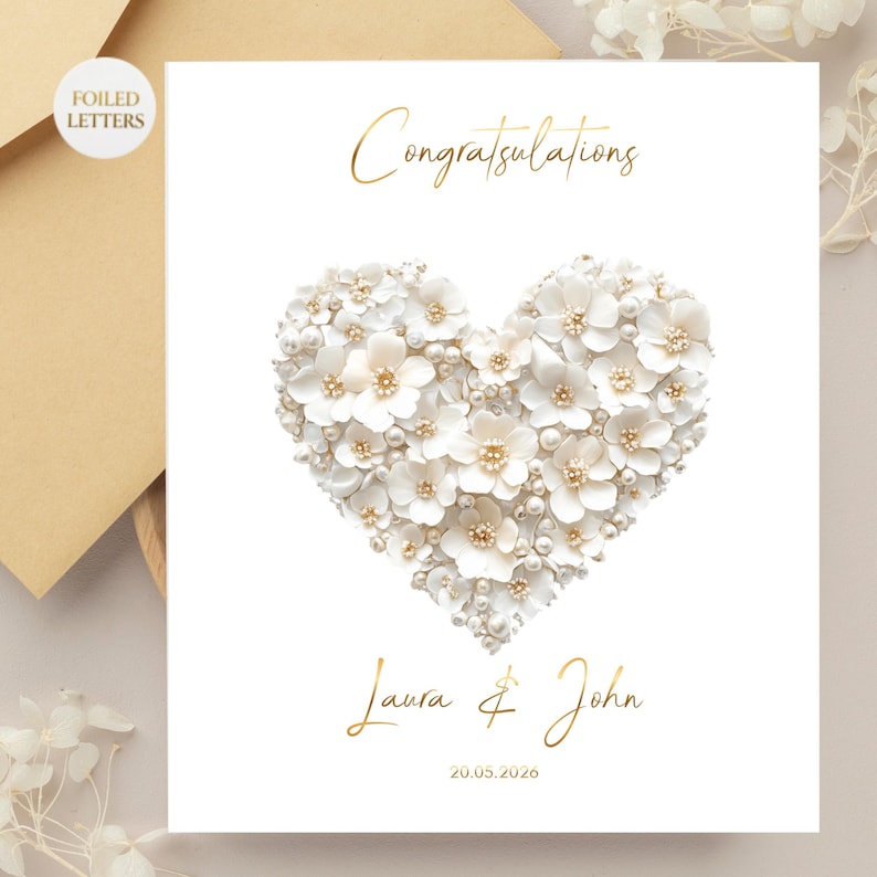 A white greeting card with a heart shape made of small white flowers and pearls, with gold foil text reading "Congratulations" above and "Laura & John 20.05.2026" below, on a light brown envelope and a blurred background.
