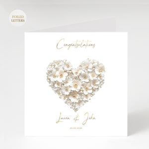 A white greeting card with a heart shape made of small white flowers and pearls, with gold foil text reading "Congratulations" above and "Laura & John 20.05.2026" below.