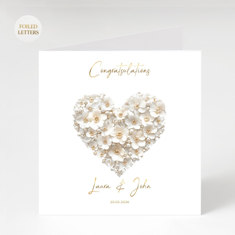 A white greeting card with a heart shape made of small white flowers and pearls, with gold foil text reading "Congratulations" above and "Laura & John 20.05.2026" below.