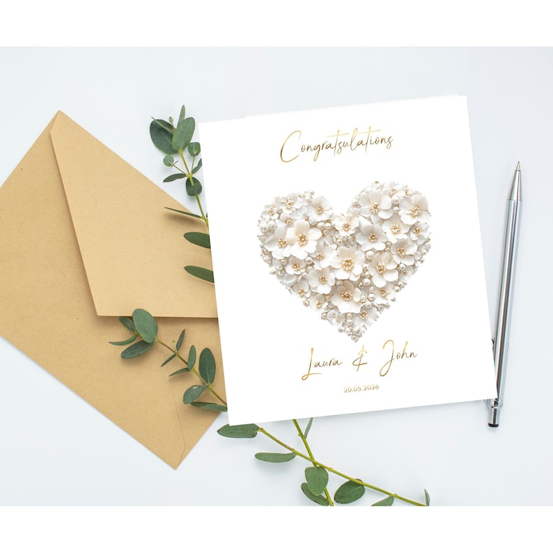 A white greeting card with a heart shape made of small white flowers and pearls, with gold foil text, next to a brown envelope, a pen, and green foliage.