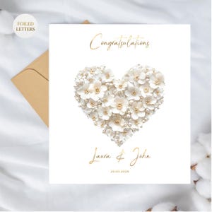 A white greeting card with a heart shape made of small white flowers and pearls, with gold foil text, on a white fabric surface with a brown envelope and cotton plant.