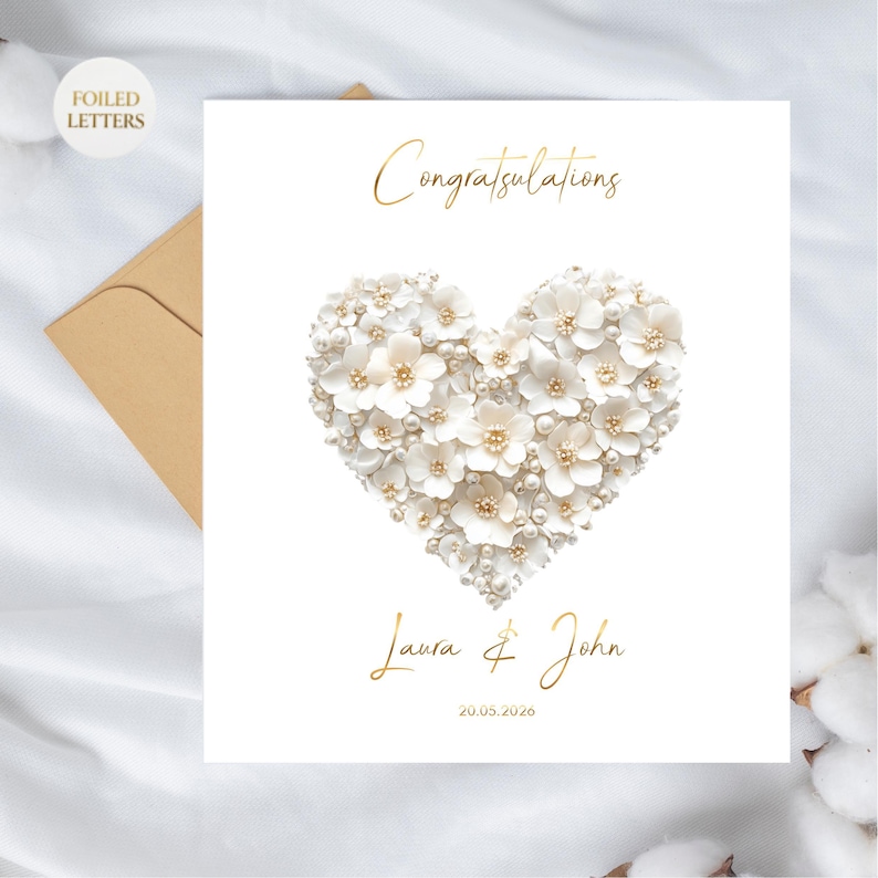A white greeting card with a heart shape made of small white flowers and pearls, with gold foil text, on a white fabric surface with a brown envelope and cotton plant.