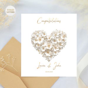 A white greeting card with a heart shape made of small white flowers and pearls, with gold foil text, on a brown envelope with dried white flowers.