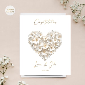 A white greeting card with a heart shape made of small white flowers and pearls, with gold foil text, on a white surface with white baby's breath flowers.