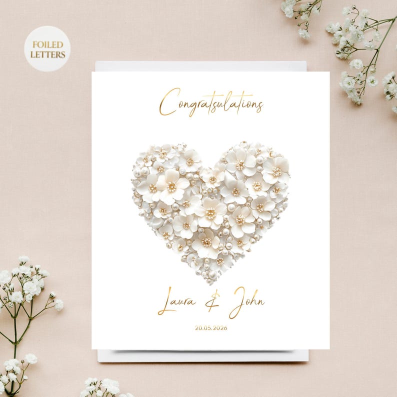 A white greeting card with a heart shape made of small white flowers and pearls, with gold foil text, on a white surface with white baby's breath flowers.