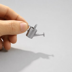 May include: A miniature silver-colored watering can with a cylindrical body, a thin handle, and a narrow spout. The small object is held between fingers, showcasing its tiny scale and metallic finish, with a clear shadow cast below it.