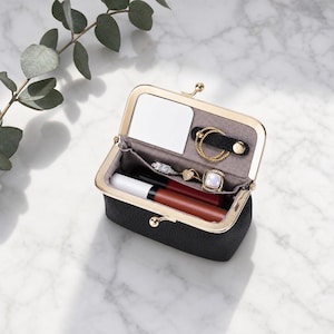 May include: A black textured leatherette cosmetic pouch with a gold-toned metal frame and kiss-lock closure. The interior features a small rectangular mirror, a snap-button ring holder, and a compartment containing two lipsticks and three rings.