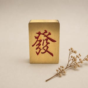 Könnte beinhalten: Ein rechteckiger, goldfarbener Mahjong-Spielstein mit einem tiefroten, eingravierten chinesischen Schriftzeichen für "Fa" oder "Glück" auf der Vorderseite. Der Spielstein hat eine glatte, metallische Oberfläche und steht aufrecht neben einem kleinen, getrockneten weißen Blumenzweig.