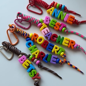 May include: Four lanyards with carabiners, each featuring colourful plastic letter beads spelling out the names Annie, Best Teacher, Robert, Charlie, and Betsy. The lanyards are made of braided cord in various patterns and colours, including red, pink, orange, and brown.