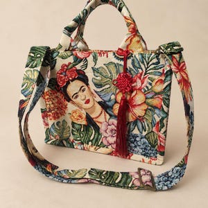 Op de afbeelding: Een rechthoekige draagtas van geweven stof met een kleurrijk bloemenpatroon en een portret van Frida Kahlo. De tas heeft twee handvatten, een verstelbare schouderriem en een decoratief rood geknoopt kwastje dat aan de voorkant hangt.