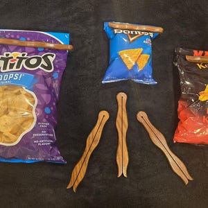 Puede incluir: Se muestran tres clips de madera para bolsas, uno asegurando una bolsa grande de Tostitos Scoops, uno en una bolsa pequeña de Doritos y otro en una bolsa de patatas fritas Martin's Honey BBQ Kettle Cook'd. Tres clips más están debajo.