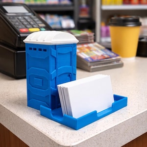 May include: A blue miniature portable toilet-shaped business card holder with a white roof. The holder has a slot to hold a stack of white business cards. The holder is on a countertop.