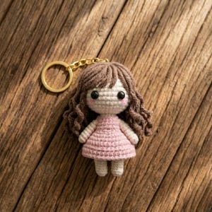 May include: A handmade crochet doll keychain. The doll has brown curly hair, rosy cheeks, and a pink dress. The keychain includes a gold-coloured ring and chain. The doll is approximately 7.6 cm tall.