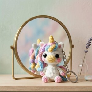 May include: A crocheted unicorn keychain with a rainbow mane, tail, and horn. The white unicorn has pink cheeks and large black eyes. It is sitting in front of a gold-framed mirror.