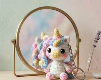 May include: A crocheted unicorn keychain with a rainbow mane, tail, and horn. The white unicorn has pink cheeks and large black eyes. It is sitting in front of a gold-framed mirror.