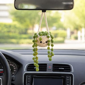 May include: A crocheted car charm featuring a smiling, beige pot with green beaded plant strands hanging down. The charm is suspended from the rearview mirror. The car's dashboard and windshield are visible in the foreground, with a blurred green background.