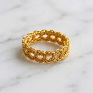 May include: A gold-colored ring with a woven design. The ring features a series of interconnected loops, creating an open, textured pattern. The ring is made of a shiny, metallic material and is set against a white marble surface.