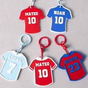 May include: Five jersey-shaped keychains with plastic clips. Two are red with white text MATEO 10, one is blue with white text NOAH 10, one is light blue with white text LUCAS 7, and one is red with blue trim and blue text OLIVER 23.