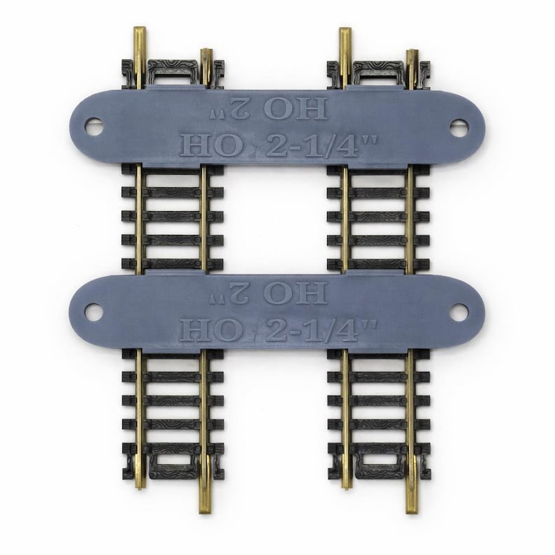 Op de afbeelding: Twee HO-schaal modeltreinbaanstukken. Elke sectie heeft zwarte rails met messingkleurige connectoren en een grijze plastic basis. De tekst "HO 2" en "HO 2-1/4" is gedrukt op de grijze basis.