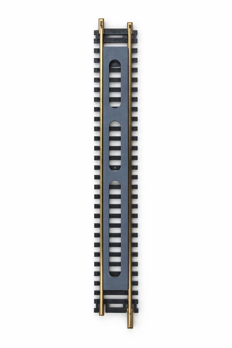 Op de afbeelding: Een sectie modeltreinrails met zwarte rails en een grijs middenstuk met drie rechthoekige uitsparingen. De rails heeft goudkleurige eindstukken, tegen een witte achtergrond.