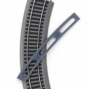 Op de afbeelding: Gebogen grijs modeltreinbaansectie met parallelle zilveren rails. Een grijze plastic meettool met "18" erop gedrukt is diagonaal over de baan geplaatst. De baan is ontworpen voor modelspoorwegen.