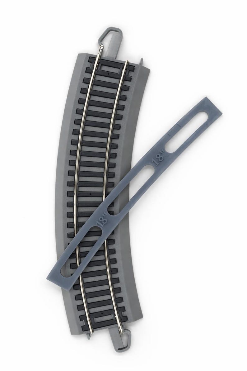 Op de afbeelding: Gebogen grijs modeltreinbaansectie met parallelle zilveren rails. Een grijze plastic meettool met "18" erop gedrukt is diagonaal over de baan geplaatst. De baan is ontworpen voor modelspoorwegen.