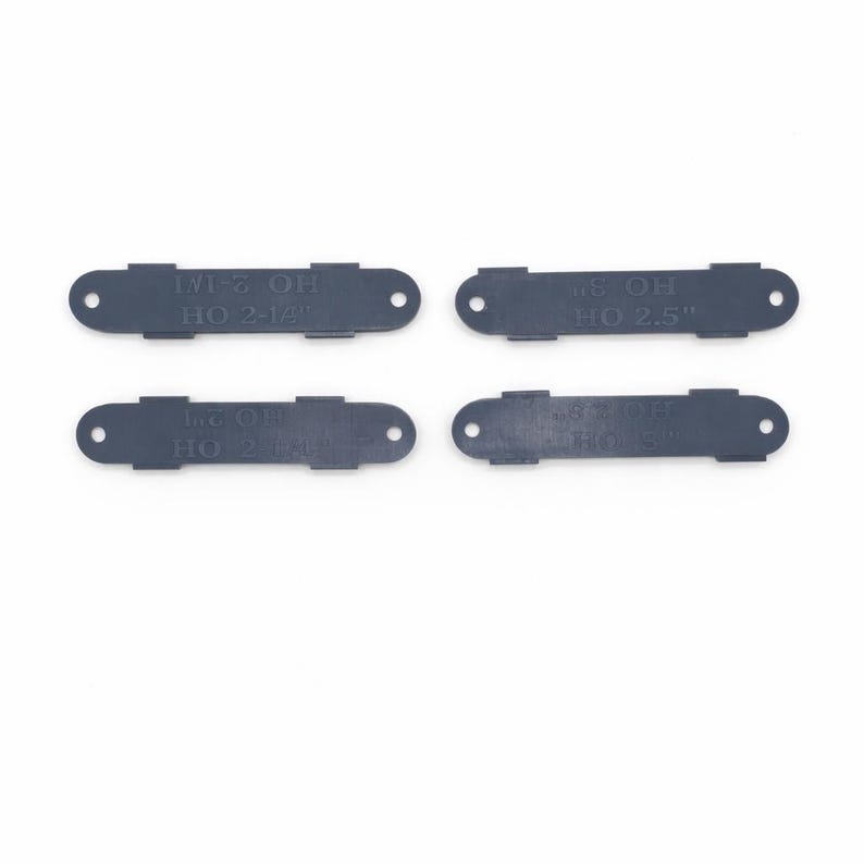 Op de afbeelding: Vier donkergrijze rechthoekige plastic stukken met afgeronde uiteinden en tekst. Elk stuk heeft twee gaten en een verhoogd middengedeelte. De tekst bevat "1/2 OH HO 2-1/4", "3/8 OH HO 2.5" en vergelijkbare markeringen.
