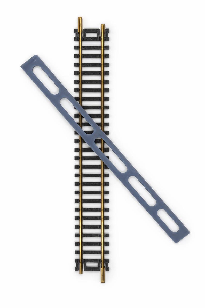 Op de afbeelding: Een close-up van modeltreinrails met een grijze plastic spooruitlijningsgereedschap. De rails zijn zwart met goudkleurige rails. Het uitlijningsgereedschap is een lang, dun stuk met twee grote ovale uitsparingen.