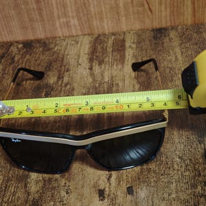 Peut inclure: Lunettes de soleil noires avec des accents dor&eacute;s et un logo Ray-Ban. Les lunettes mesurent environ 14 cm de largeur. Les montures sont noires avec une bordure dor&eacute;e sur le bord sup&eacute;rieur. Les branches sont fines et dor&eacute;es.