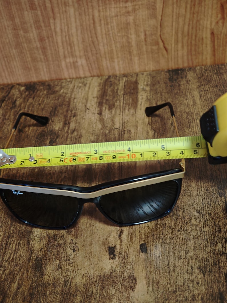 Peut inclure: Lunettes de soleil noires avec des accents dor&eacute;s et un logo Ray-Ban. Les lunettes mesurent environ 14 cm de largeur. Les montures sont noires avec une bordure dor&eacute;e sur le bord sup&eacute;rieur. Les branches sont fines et dor&eacute;es.