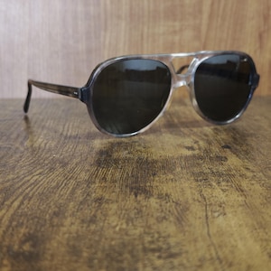 Puede incluir: Gafas de sol con montura transparente y lentes oscuras. Las gafas tienen un doble puente y una forma de aviador clásica. Las patillas son de color marrón oscuro. Las gafas descansan sobre una superficie de madera.