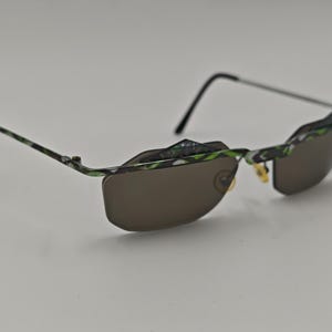 Puede incluir: Gafas de sol con un patrón de camuflaje en las monturas. Las lentes son de color marrón oscuro y las monturas tienen un patrón verde, negro y blanco. Las patillas son negras.