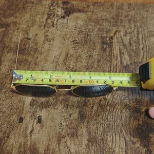 May include: Gold-framed sunglasses with dark lenses are measured with a yellow tape measure. The sunglasses are approximately 5 inches wide. The background is a wooden surface.